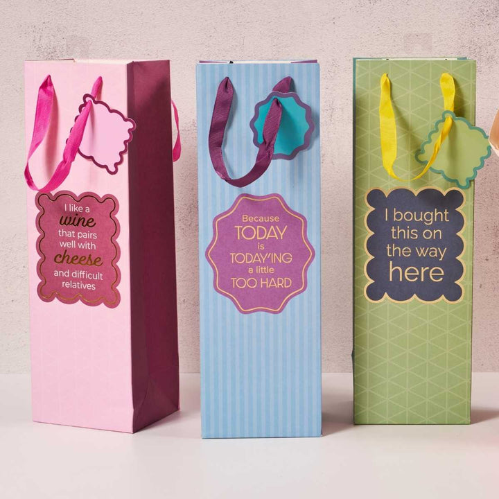Drinks On Me’s Today'ing-Wine Bag set includes three reusable bags with ribbon handles and witty sayings—“wine and cheese” (pink), “today is too hard” (blue), and “I bought this on the way here” (green)—perfect hostess gifts.