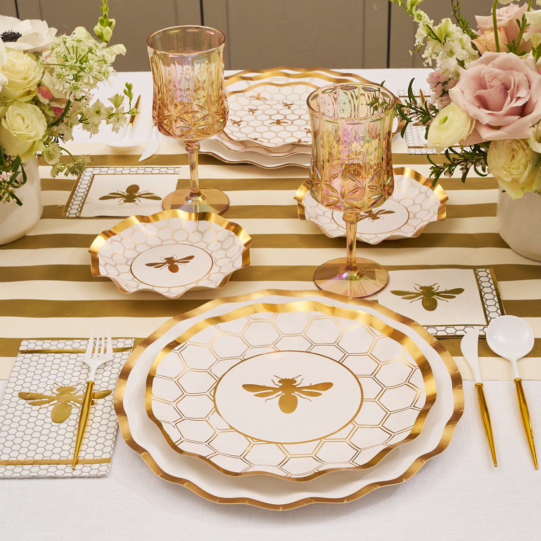 A table set with Sophistiplate Honeybee White Wavy Paper Dinner Plates (8pk), gold-rimmed disposable glasses, bee and honeycomb decor, gold cutlery, white napkins, a gold bee centerpiece, and a gold-striped runner.