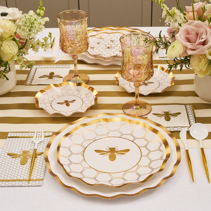 A table set with Sophistiplate Honeybee White Wavy Paper Dinner Plates (8pk), gold-rimmed disposable glasses, bee and honeycomb decor, gold cutlery, white napkins, a gold bee centerpiece, and a gold-striped runner.