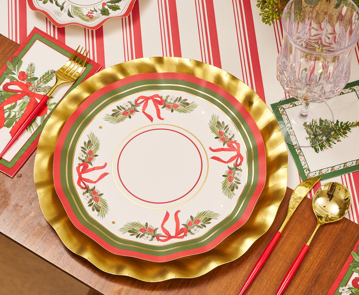 A festive table setting featuring Sophistiplate’s Home for the Holidays Dinner Plates (8pk), adorned with gold rims and charming red and green holiday designs, paired with elegant gold utensils, a crystal glass, and cheerful holiday-themed napkins on a vibrant red and white striped tablecloth—the perfect way to showcase your Sophistiplate Christmas party or dinner plates in style.