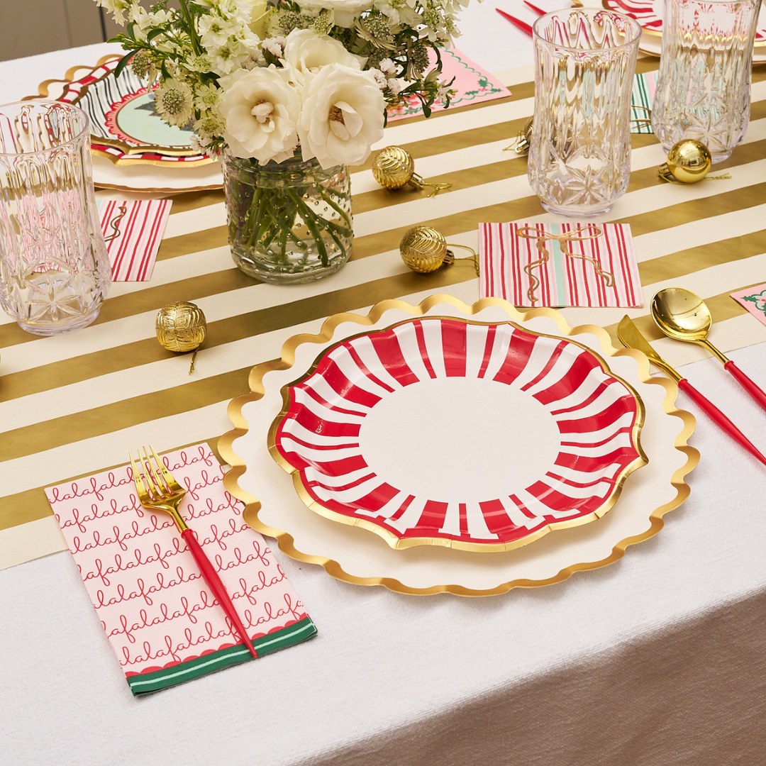 Sophistiplate “Tis the Season” Dinner Plates (8-pack) create a festive table setting when paired with gold utensils, crystal glasses, a vase of white flowers, gold ornaments, and a white tablecloth with a gold-striped runner.
