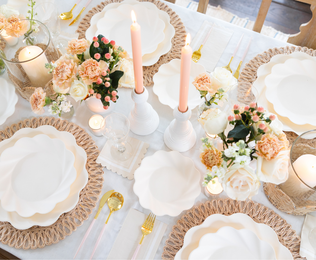 The Eco-Friendly Table Setting-Cream by Sophistiplate features scalloped white compostable plates, gold cutlery, pink candles, and floral centerpieces with white and peach blooms on a white tablecloth for a warm, inviting tablescape.