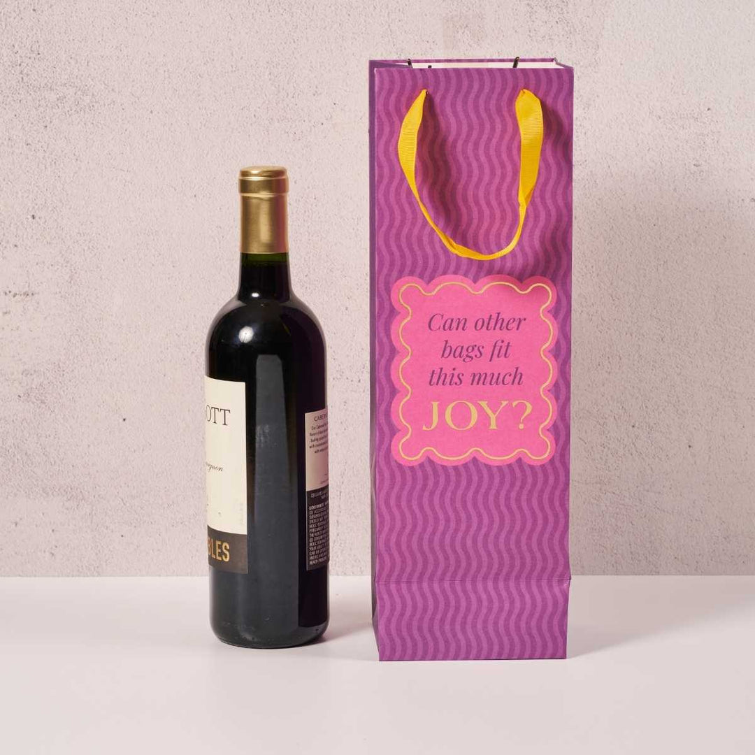 A bottle of red wine stands next to the Drinks On Me Joy Bag, a funny wine bag with yellow handles and a pink label that says, “Can other bags fit this much JOY?” on a light background.