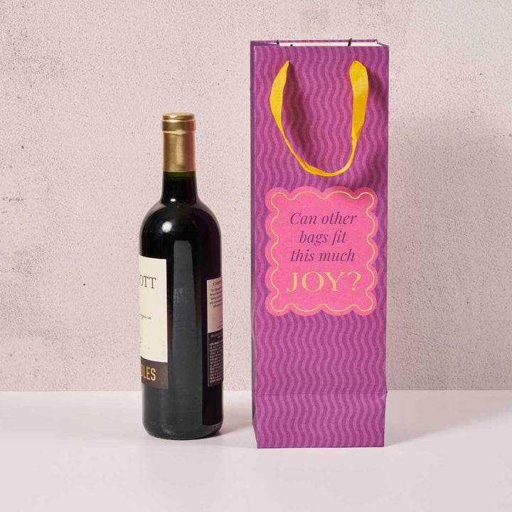 A bottle of red wine stands next to the Drinks On Me Joy Bag, a funny wine bag with yellow handles and a pink label that says, “Can other bags fit this much JOY?” on a light background.