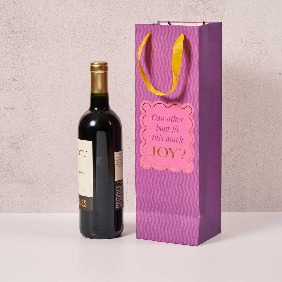 A bottle of wine stands beside the Joy Bag- Wine Bag by Drinks On Me, featuring yellow handles and a label that asks, "Can other bags fit this much JOY?" on a light surface against a neutral background.
