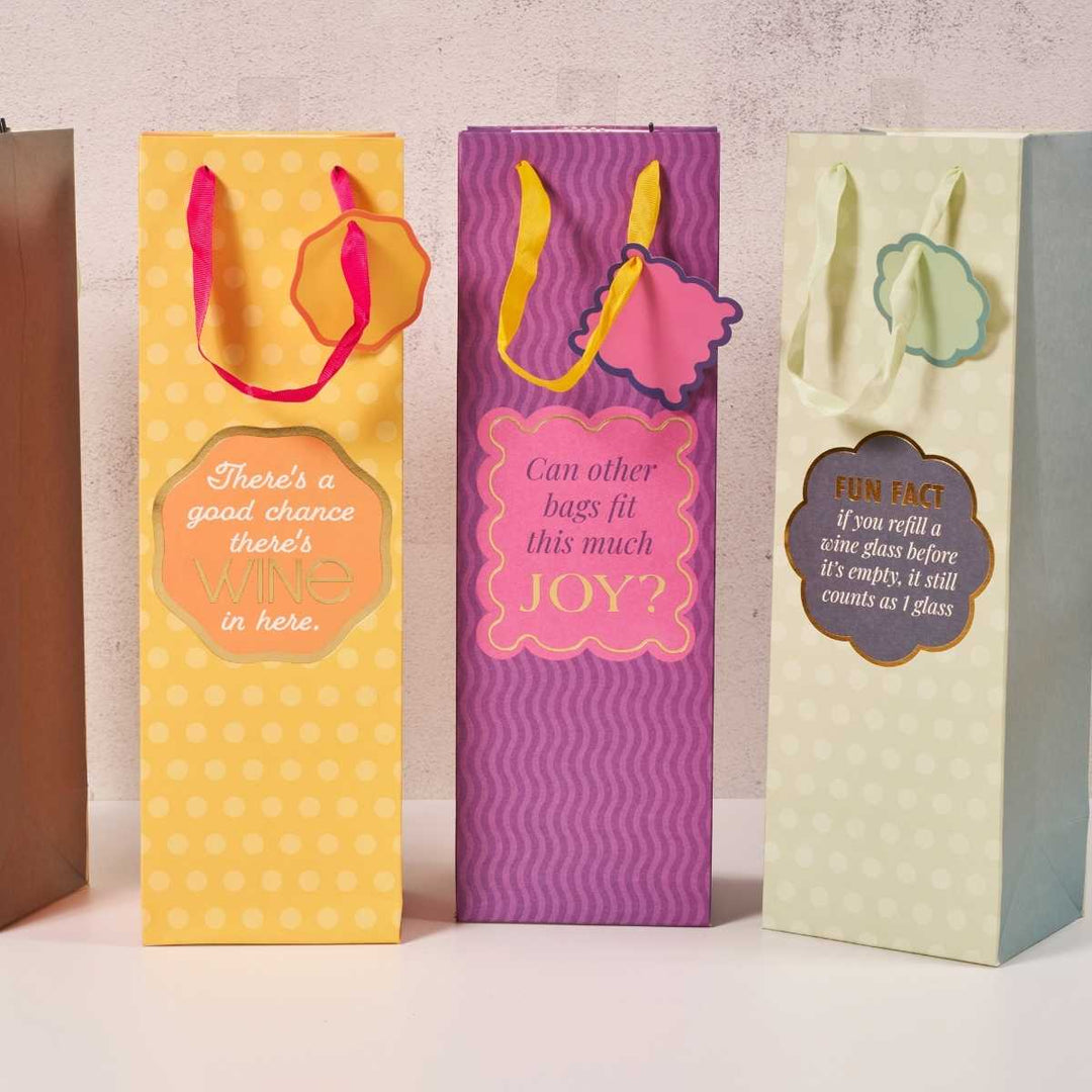 Three Joy Bag-Wine Bags by Drinks On Me, featuring tall reusable designs with colorful patterns, humorous wine phrases, and matching ribbon handles, stand upright side by side against a light background.