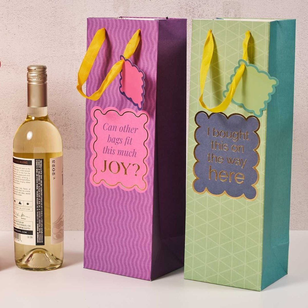 A bottle of white wine stands beside two colorful Drinks On Me Joy Bag-Wine Bags with yellow handles—one reads “Can other bags fit this much JOY?” and the other says, “I bought this on the way here.”.