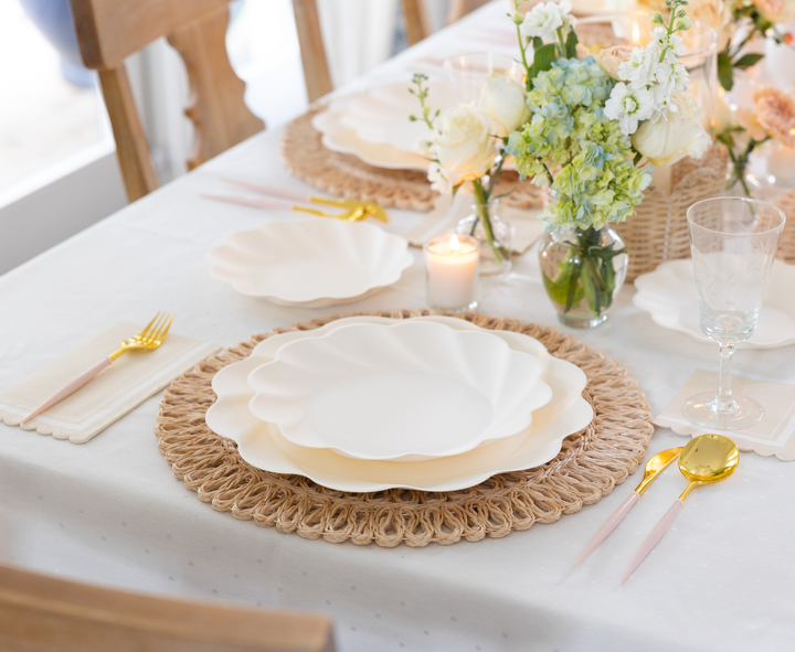 A table set for a meal with Sophistiplate Simply Eco Extra Large Plates in cream, gold and pink cutlery, woven placemats, a lit candle, and pastel floral centerpieces on a white tablecloth.