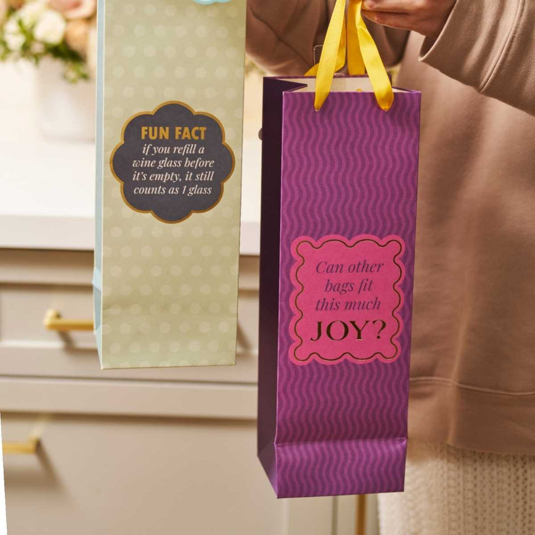 Two Drinks On Me wine gift bags: a funny bag labeled "FUN FACT: if you refill a wine glass before it’s empty, it still counts as 1 glass" and the reusable Joy Bag- Wine Bag asking, "Can other bags fit this much JOY?.