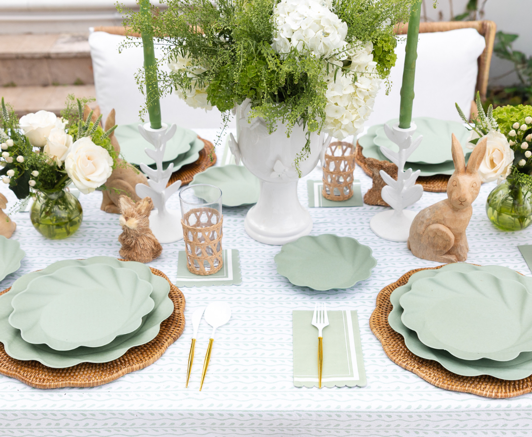 A spring table features sustainable tableware, wicker placemats, gold cutlery, green candles, white flowers, decorative wooden bunnies, and Sophistiplate's Guest Towel Scalloped Edge Sage napkins on a pale patterned tablecloth.