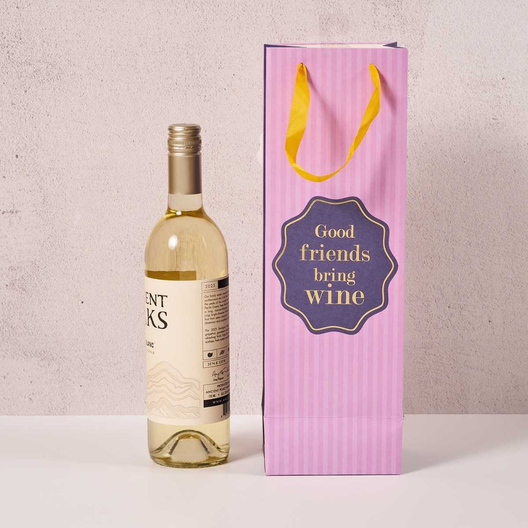 A bottle of white wine sits beside the Good Friends- Wine Bag by Drinks On Me, featuring yellow handles and "Good friends bring wine" text. The light, neutral background highlights it as a perfect hostess gift.