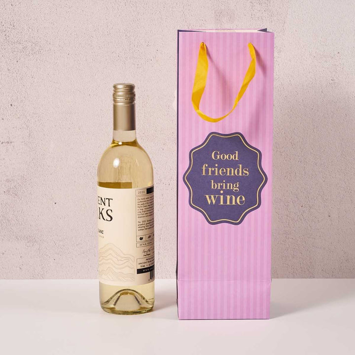 A bottle of white wine sits beside the Good Friends- Wine Bag by Drinks On Me, featuring yellow handles and "Good friends bring wine" text. The light, neutral background highlights it as a perfect hostess gift.