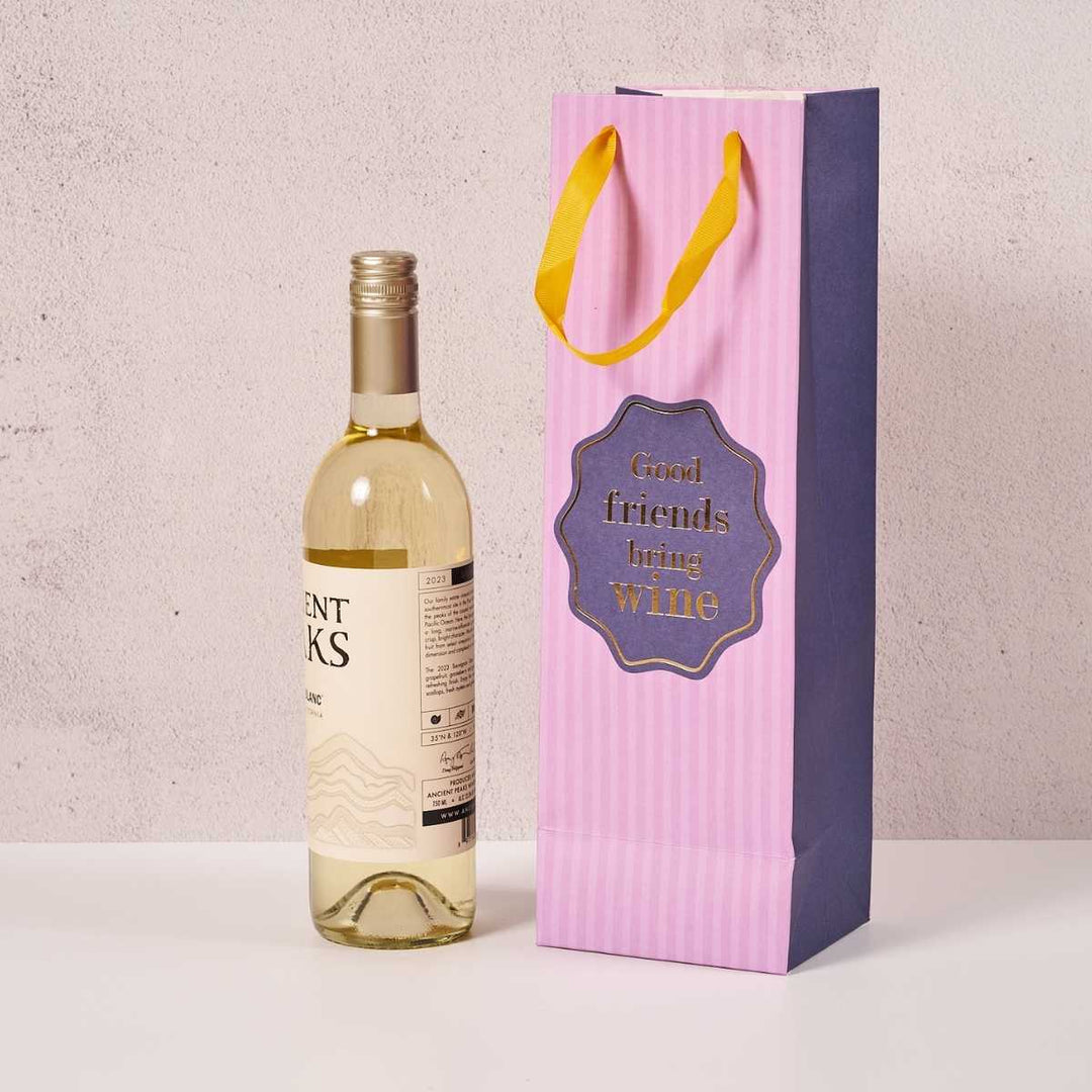 A Drinks On Me Good Friends- Wine Bag in pink and purple with yellow handles stands beside a white wine bottle—the bag reads "Good friends bring wine," making it a perfect reusable gift for any celebration.