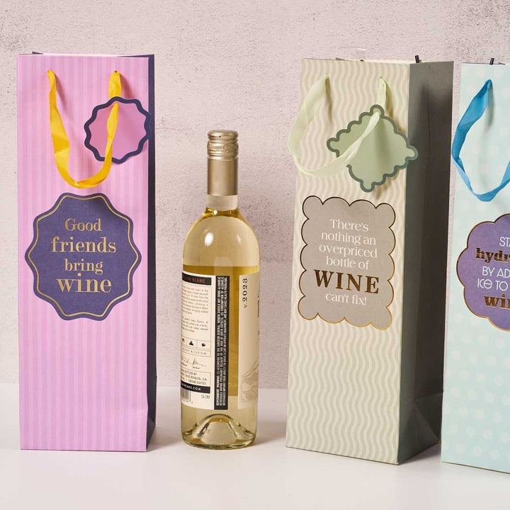 A bottle of white wine stands among three Drinks On Me "Good Friends" reusable wine bags, each with ribbon handles and a funny quote—ideal as a humorous wine bag or thoughtful hostess gift.