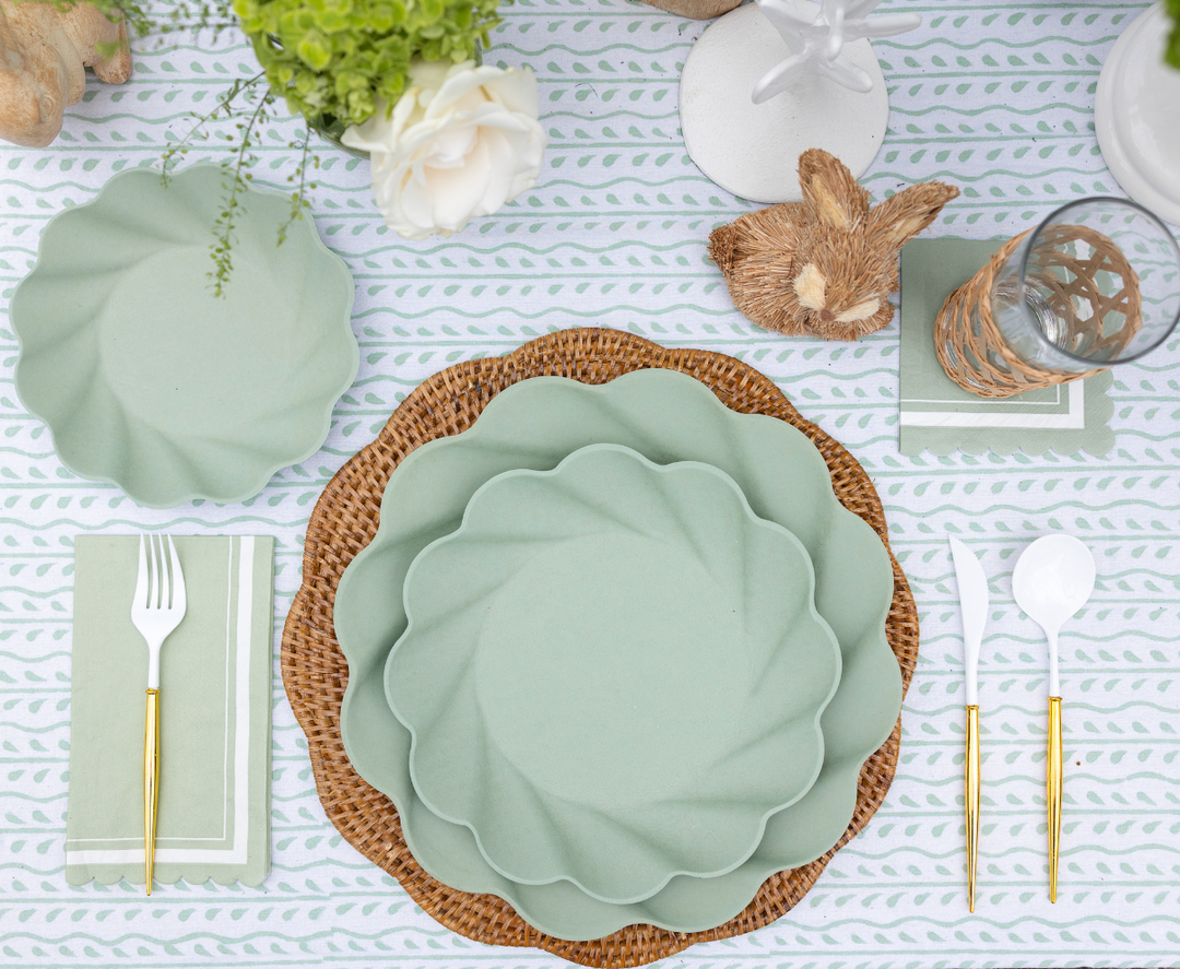 Create a spring-themed table with Sophistiplate Guest Towel Scalloped Edge Sage, eco-friendly tableware, gold-handled white cutlery on green napkins, woven placemats, a floral centerpiece, glassware, a bunny accent, and a patterned cloth.