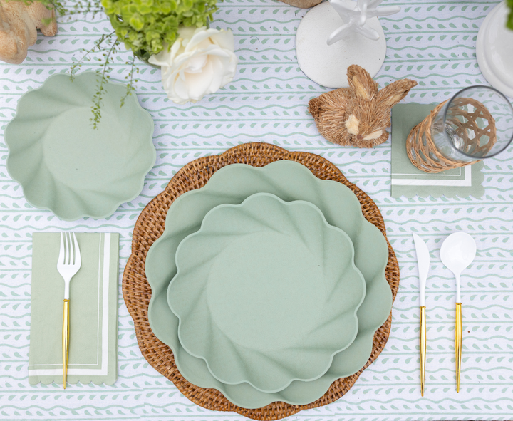 Create a spring-themed table with Sophistiplate Guest Towel Scalloped Edge Sage, eco-friendly tableware, gold-handled white cutlery on green napkins, woven placemats, a floral centerpiece, glassware, a bunny accent, and a patterned cloth.
