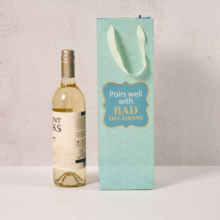 A bottle of white wine stands beside the Drinks On Me Bad Decisions Wine Bag, a light blue reusable bag with a geometric pattern and "Pairs well with BAD DECISIONS"—a funny, stylish hostess gift.