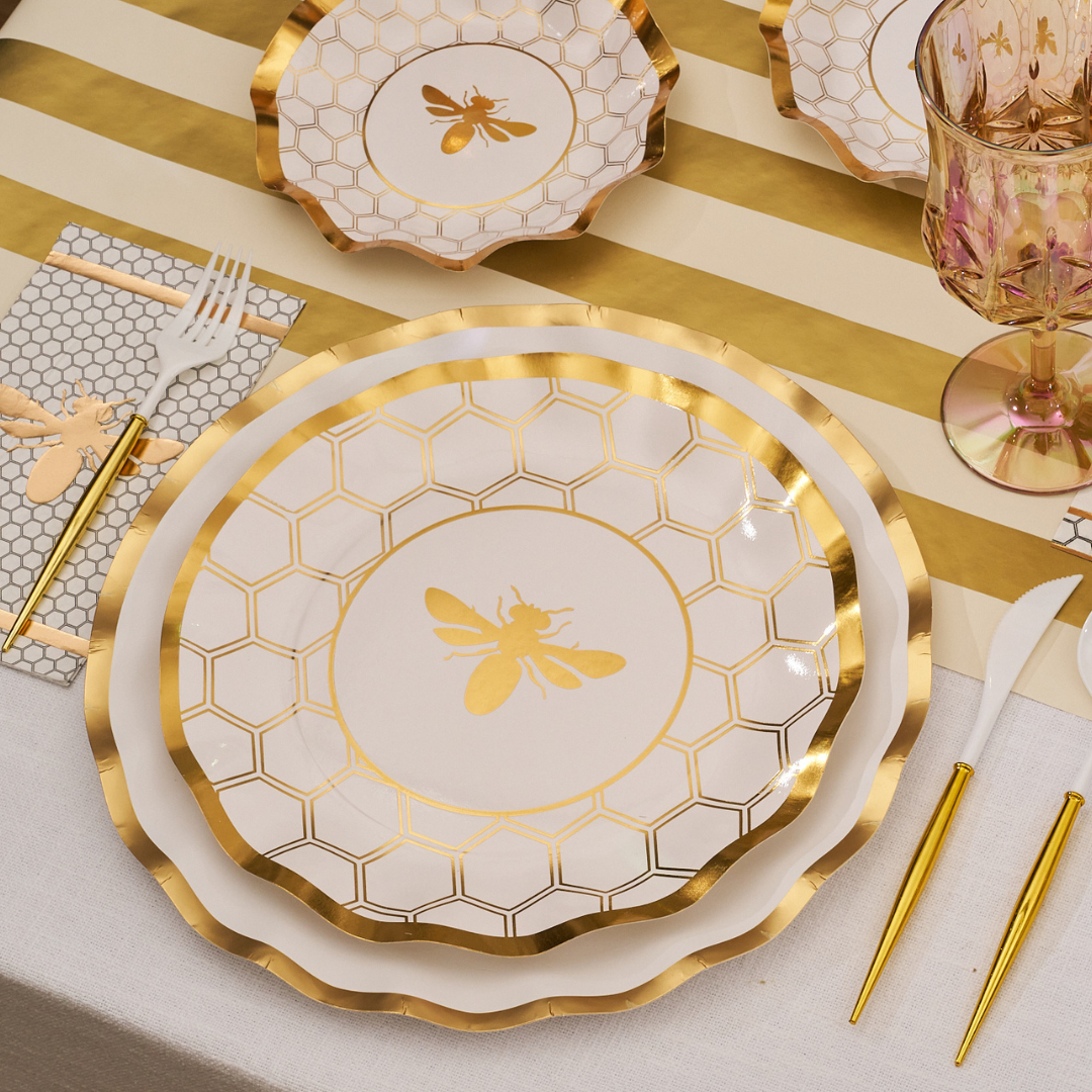 Sophistiplate's Honeybee White Wavy Paper Dinner Plate (8pk) stars in a gold and white table setting with gold bee details, gold utensils, honeycomb napkins, a pink glass, and a striped table runner.