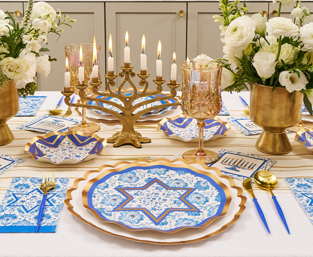 Create a festive tablescape with the Sophistiplate Hanukkah Toile Table Setting, featuring designer blue and gold disposable plates adorned with the Star of David, a menorah with glowing candles, elegant gold utensils, decorative napkins, and fresh white flower arrangements.