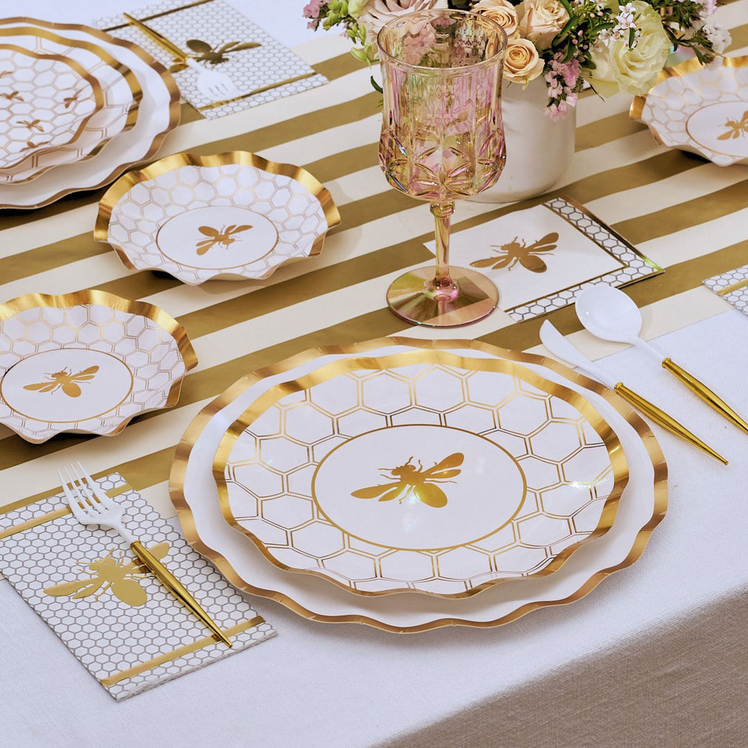 Set the scene with Sophistiplate’s White Honeybee Table Setting, featuring elegant gold and white bee-themed plates—including stylish gold rim paper and disposable plates—paired with gold utensils, patterned napkins, a pink wine glass, and a beautiful flower arrangement atop a striped tablecloth.