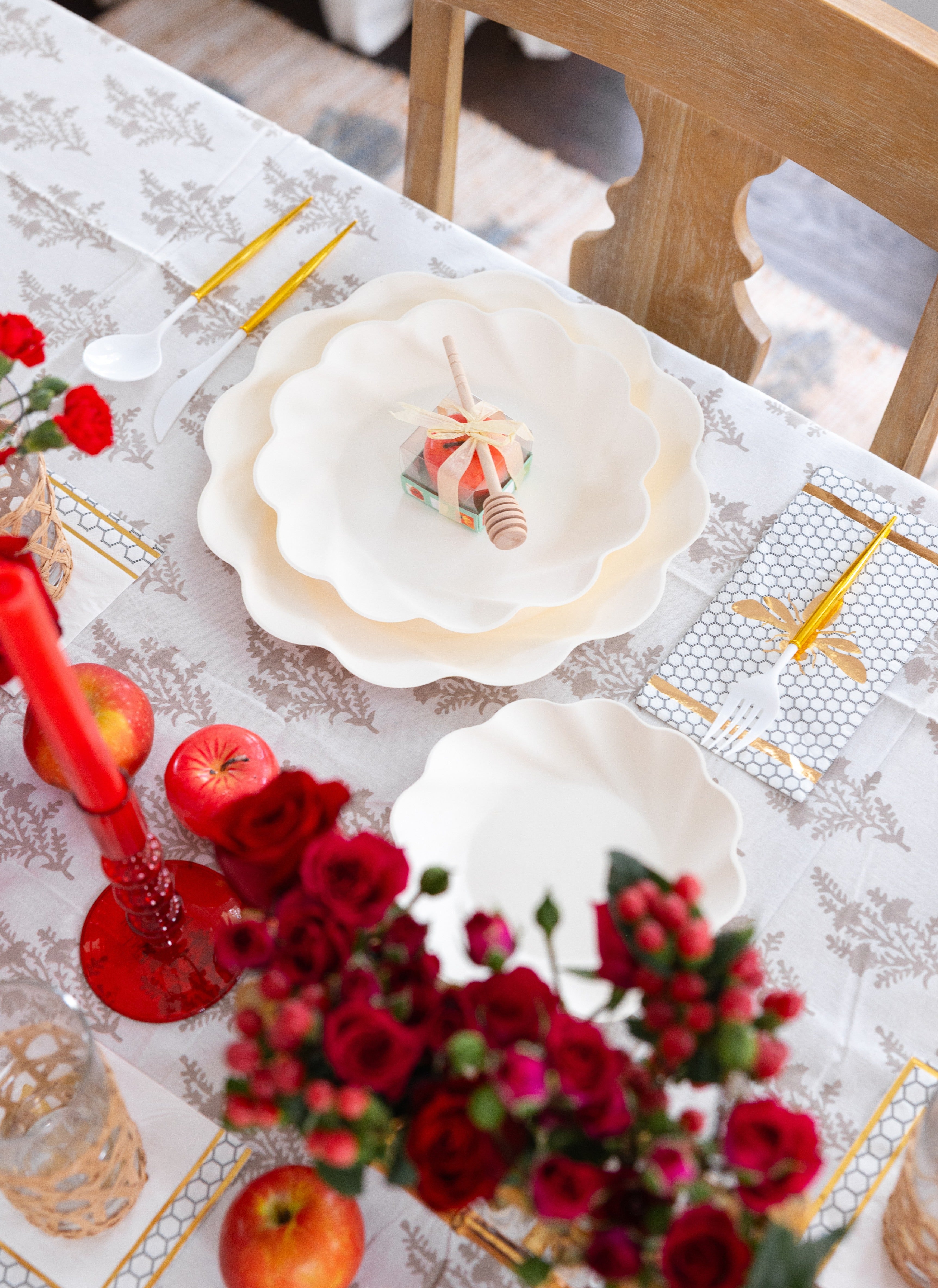 Set a festive table for Rosh Hashanah with the Sophistiplate LLC “Rosh Tablescape for 16 Guests: Eco Cream Honeybee” set, featuring scalloped white plates, gold utensils, apples, red candles, roses, and a small honey jar with dipper—all beautifully arranged on a patterned tablecloth.