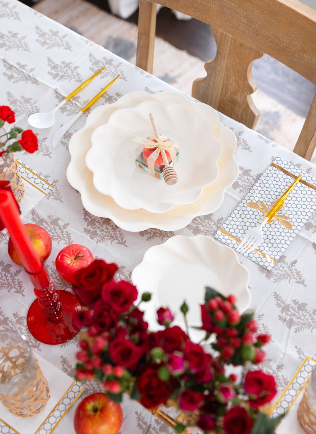 Set a festive table for Rosh Hashanah with the Sophistiplate LLC “Rosh Tablescape for 16 Guests: Eco Cream Honeybee” set, featuring scalloped white plates, gold utensils, apples, red candles, roses, and a small honey jar with dipper—all beautifully arranged on a patterned tablecloth.