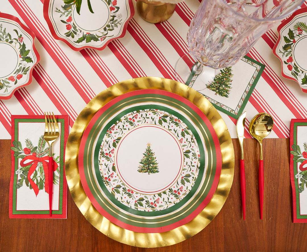 Festive table setting with a gold, red, and green Christmas tree plate, gold cutlery with red handles, a crystal glass, and Sophistiplate Home for the Holidays Guest Towels (20pk) napkins on a red and white striped table runner.