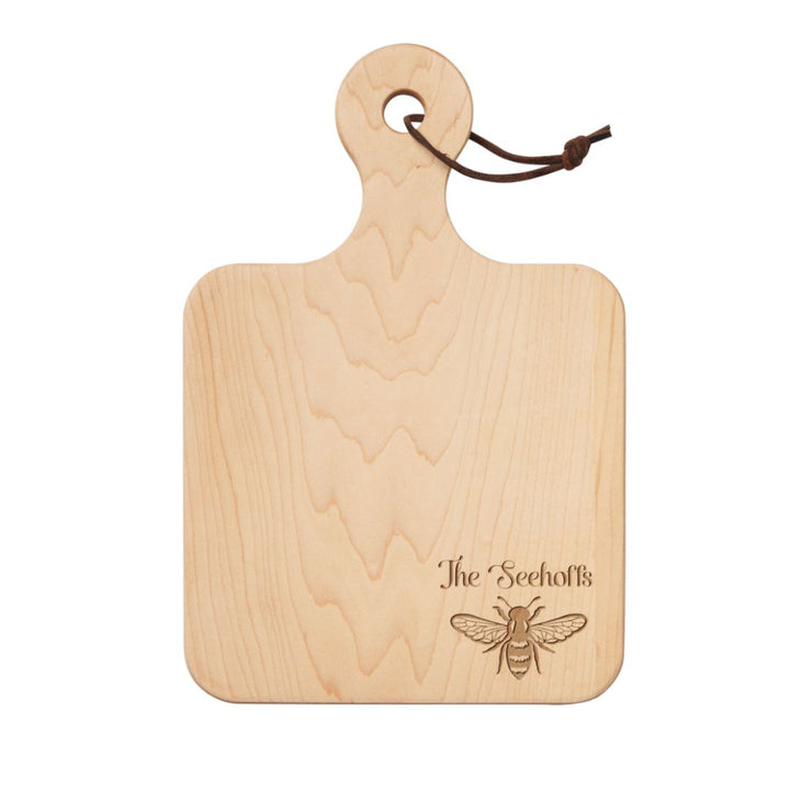 The Maple 12" x 8" Maple Wood Board features a handle, leather loop, and “The Seehoffs” engraved above a honeybee illustration—personalized and perfect as a charcuterie board for any gathering.