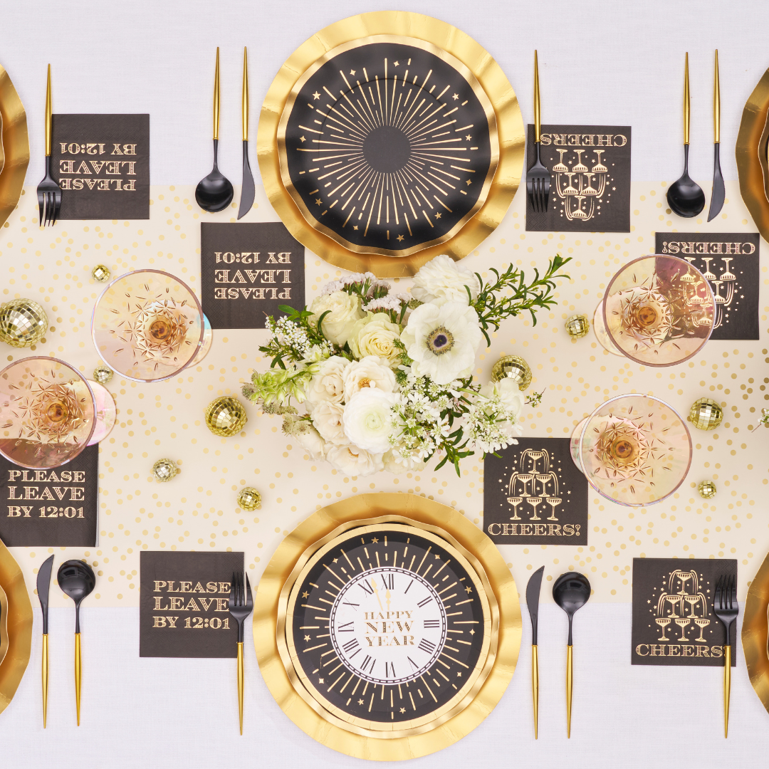 Overhead view of two place settings with Sophistiplate Midnight Sparkle Salad Plates/8pk, black cutlery, and napkins reading Please leave by 12:01 and Cheers, arranged around a floral centerpiece on a gold-polka-dot runner.