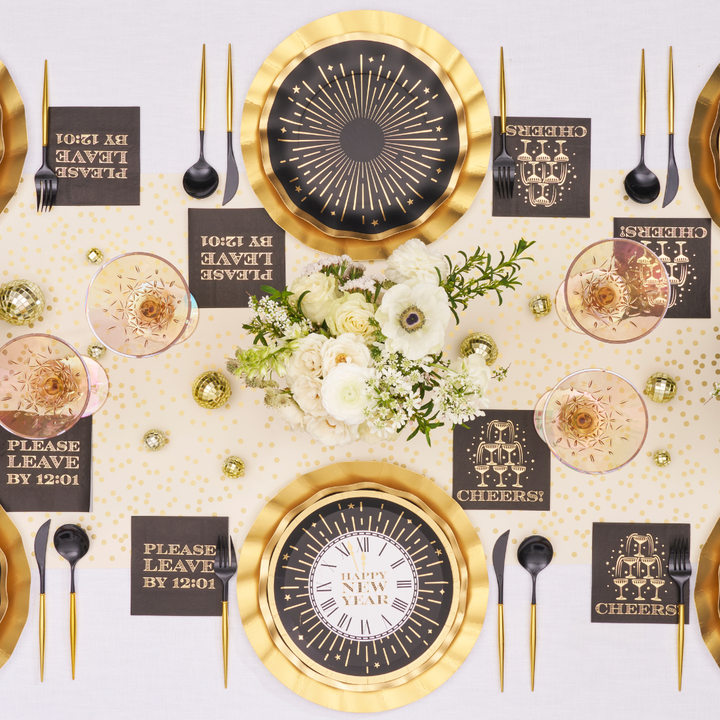 Overhead view of two place settings with Sophistiplate Midnight Sparkle Salad Plates/8pk, black cutlery, and napkins reading Please leave by 12:01 and Cheers, arranged around a floral centerpiece on a gold-polka-dot runner.