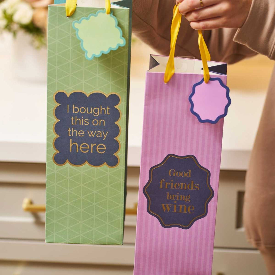 The Good Friends- Wine Bag by Drinks On Me includes two reusable bags: one green ("I bought this on the way here") and one pink ("Good friends bring wine"). Both have yellow handles—funny wine gift bags perfect for a hostess.