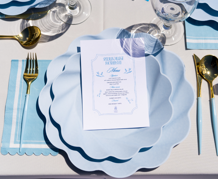 A close-up of Sophistiplate's Guest Towel Scalloped Edge Sky Blue (20/pkg) with a printed menu on top, set on compostable napkins, gold cutlery, and wine glasses atop a white tablecloth.
