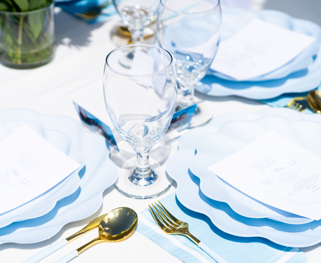 A close-up of an elegant table with Sophistiplate Simply Eco Dinner Plate- Sky Blue/8pkg, gold utensils, clear water glasses, and printed menus, beautifully arranged on a white tablecloth in bright daylight.