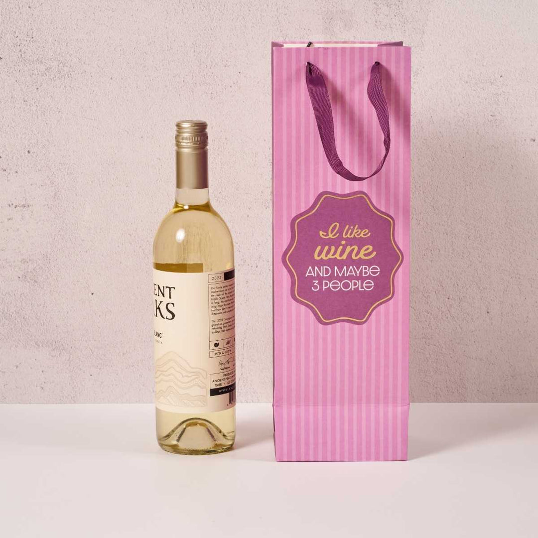 A bottle of white wine sits beside the Drinks On Me "I Like Wine" wine bag with purple handles and the quote "I like wine and maybe 3 people." Perfect as a hostess gift, both rest on a white surface against a light background.