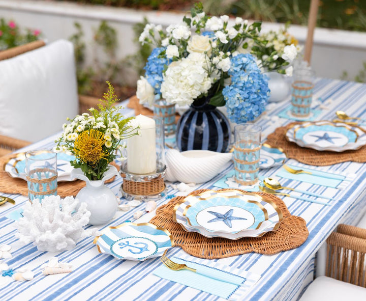 The Tidal Wave Table Setting by Sophistiplate creates a coastal party vibe with blue and white striped linens, starfish paper plates, wicker placemats, gold utensils, blue hydrangeas, and ocean-inspired accents like coral and seashells.