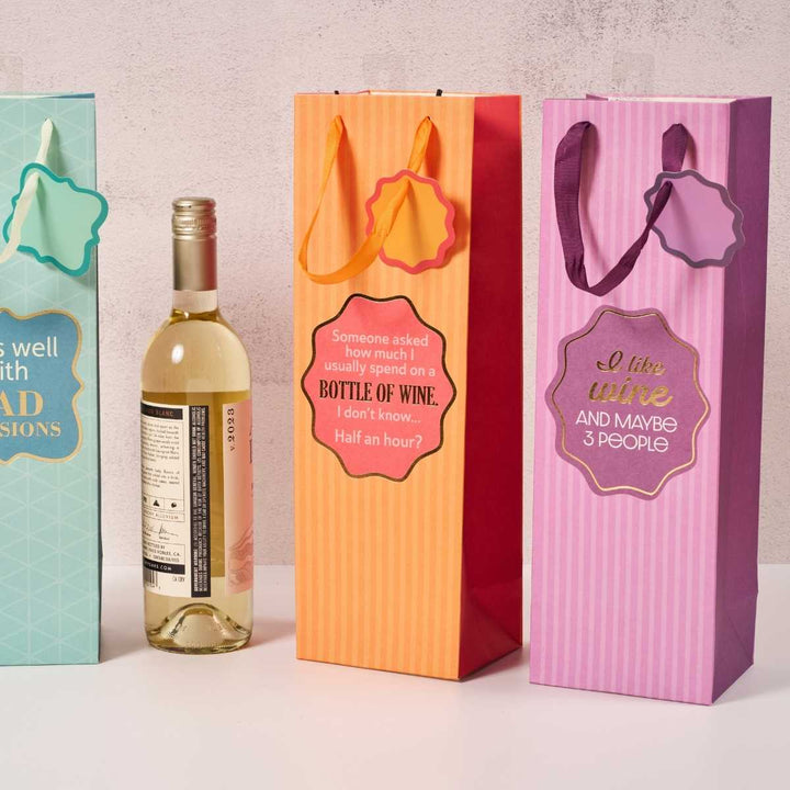 The Drinks On Me "I Like Wine" wine bags come in four colors with ribbon handles and funny sayings, shown upright beside a white wine bottle—perfect hostess gifts delivered in style.