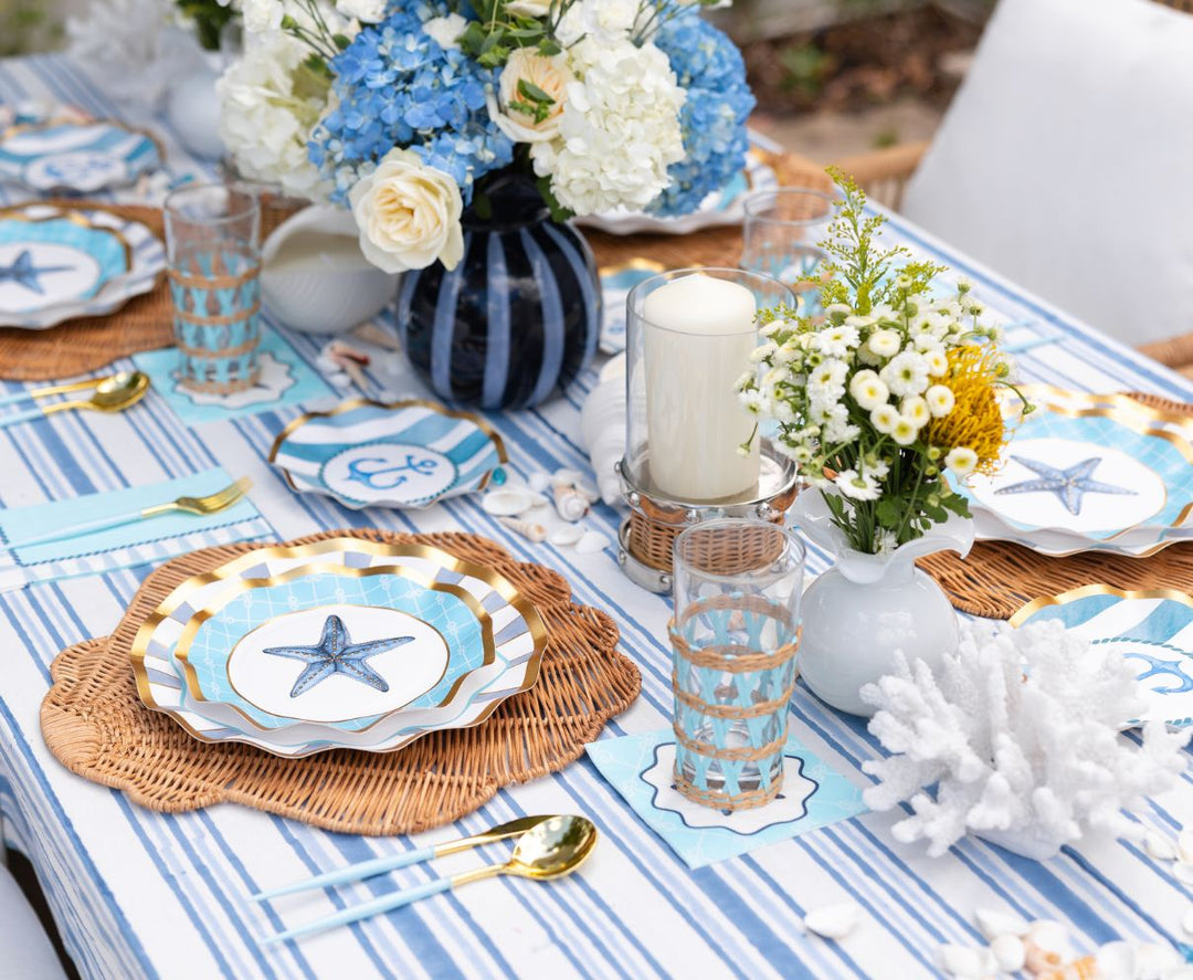 The Tidal Wave Table Setting by Sophistiplate features striped blue and white linens, starfish designer plates, woven placemats, gold cutlery, hydrangeas, white flowers, candles, and coral décor for a coastal-inspired tablescape.