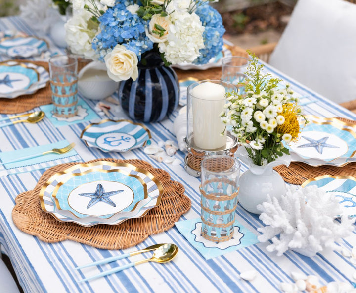 The Tidal Wave Table Setting by Sophistiplate features striped blue and white linens, starfish designer plates, woven placemats, gold cutlery, hydrangeas, white flowers, candles, and coral décor for a coastal-inspired tablescape.
