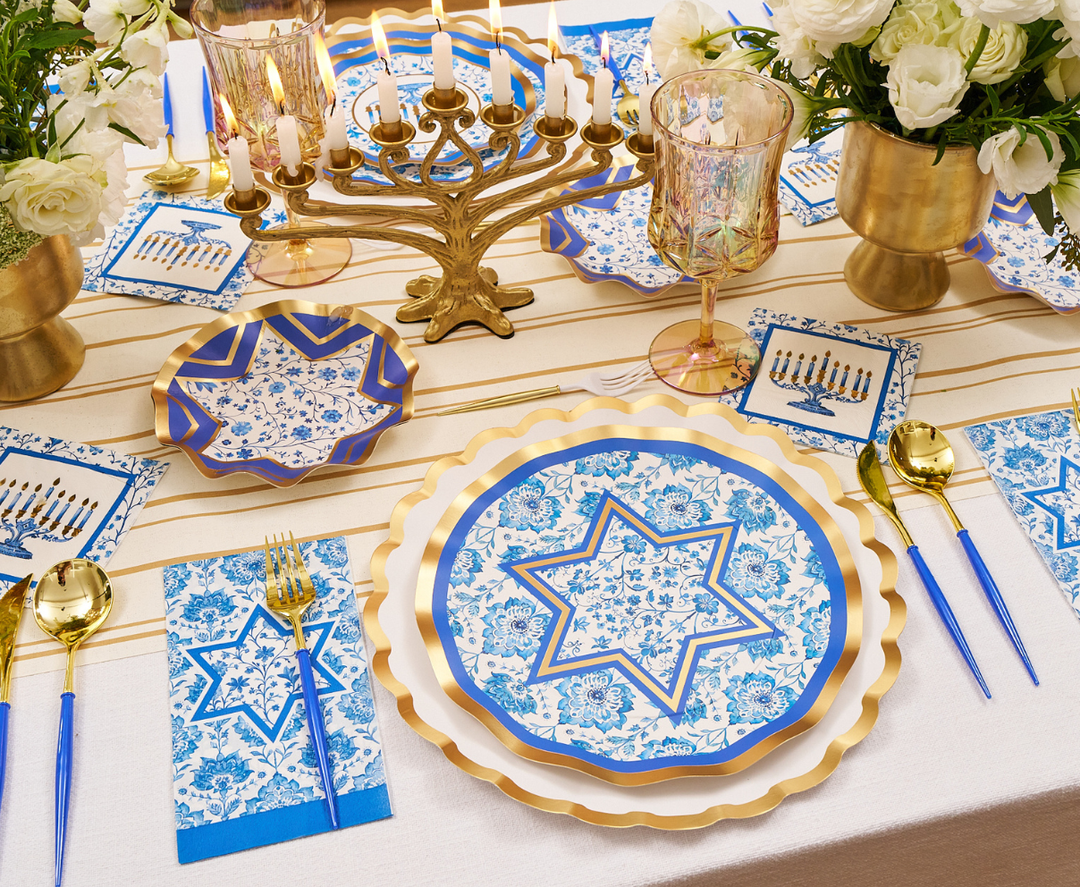 Create a truly festive tablescape with the Sophistiplate Hanukkah Toile Table Setting, featuring designer paper plates adorned with floral and Star of David motifs, elegant gold cutlery, a menorah with candles, white flowers in gold vases, festive napkins, and a white and gold striped tablecloth for an elegant Hanukkah celebration.