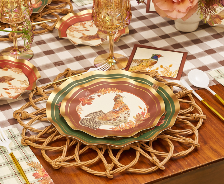 A decorative table setting with scalloped plates featuring a pheasant design and Sophistiplate Woodland Harvest Salad Plates (8pk), gold-trimmed glasses, white cutlery, and a woven placemat on a brown and white checkered tablecloth.