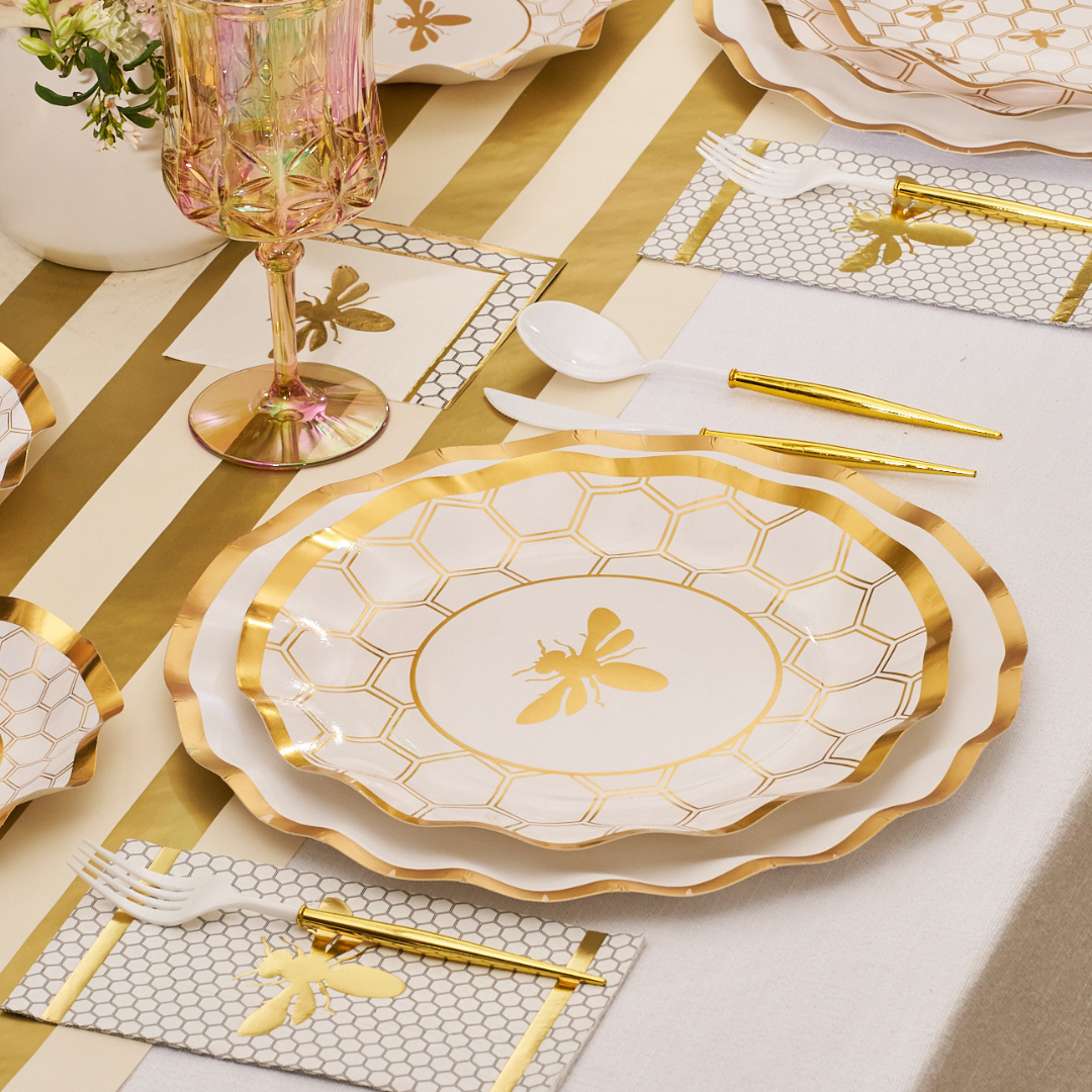 A table is set with Sophistiplate Honeybee White Wavy Paper Dinner Plates, gold cutlery, a pink glass, a bee-motif white napkin, and a gold-striped runner.