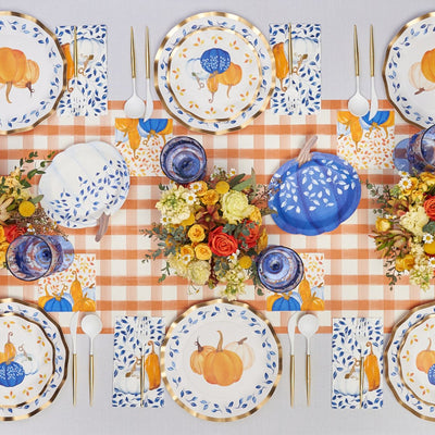 A festive autumn table is set with Sophistiplate Wavy Salad Plate Harvest Blues, blue and white napkins, gold cutlery, blue glasses, floral centerpieces, and decorative pumpkins on an orange checkered table runner.