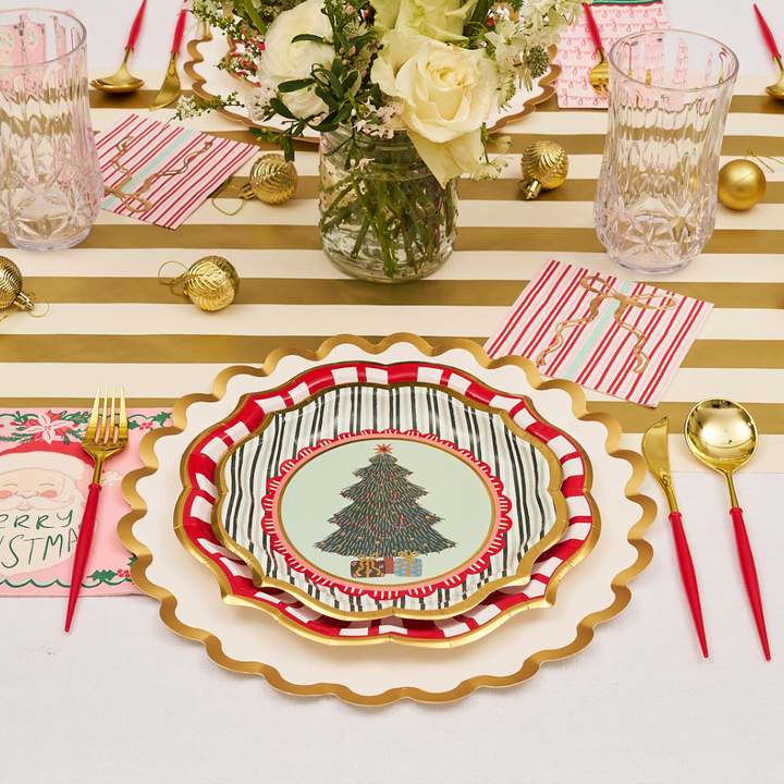Create a festive table setting with a gold-striped runner, gold-rimmed stacked plates featuring a Christmas tree, Sophistiplate Tis the Season Salad Plates (8pk), red and gold utensils, glasses, napkins, gold ornaments, and a vase of white flowers.