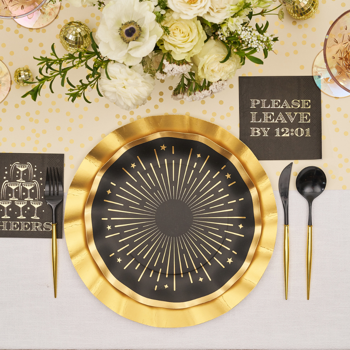 A Midnight Sparkle Table Setting featuring gold-rimmed black designer disposable plates with a starburst design, gold-handled cutlery, and black napkins printed with Cheers and Please leave by 12:01.