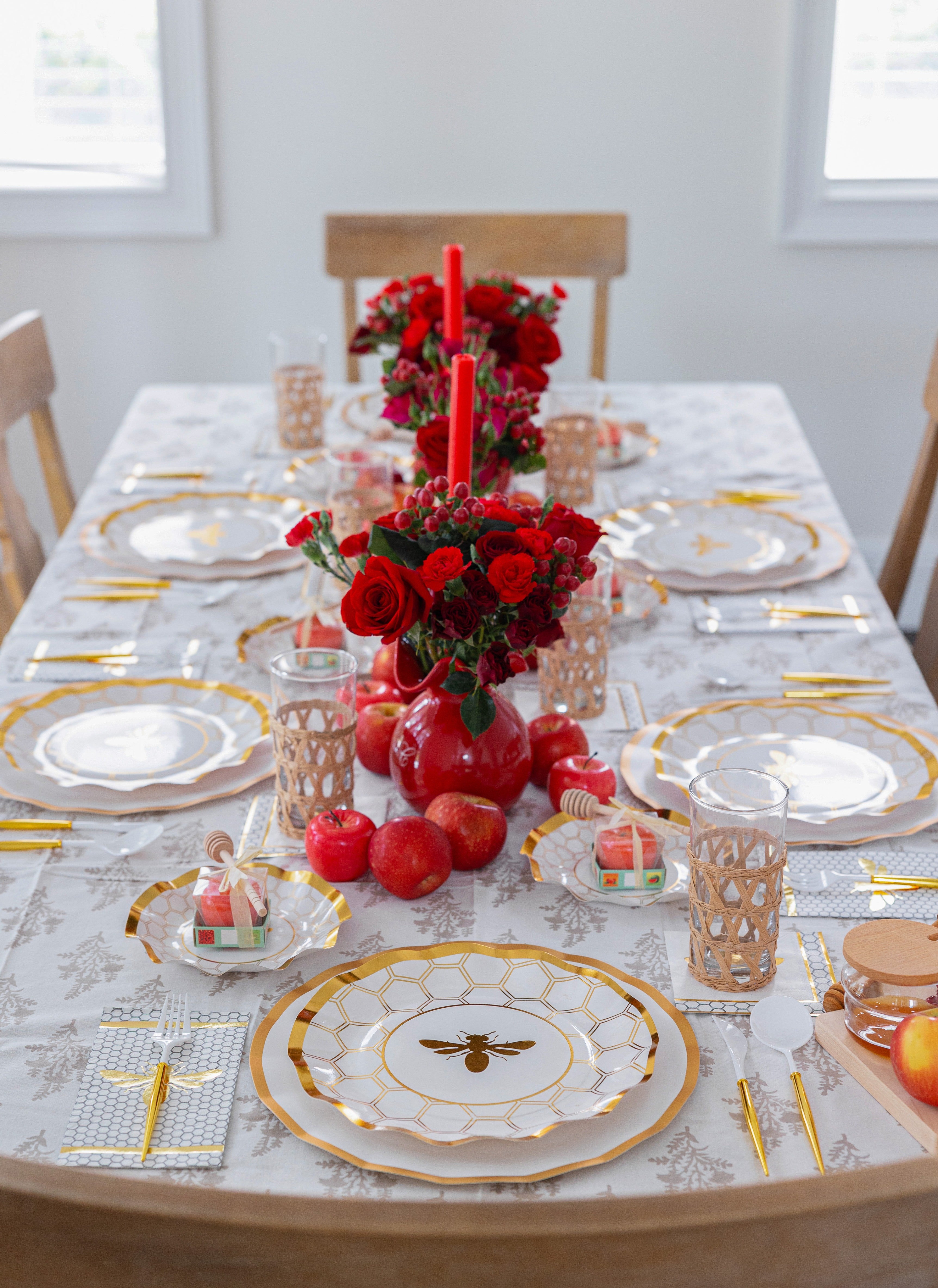 Sophistiplate LLC’s “Rosh Tablescape for 16 Guests: White Honeybee” sets a sophisticated Rosh Hashanah table with its gold-trimmed disposable plates featuring an elegant bee motif. Red apples, candles, and vases of red roses complement the gold cutlery and lattice-designed glasses, all artfully arranged around gift boxes on a patterned tablecloth.