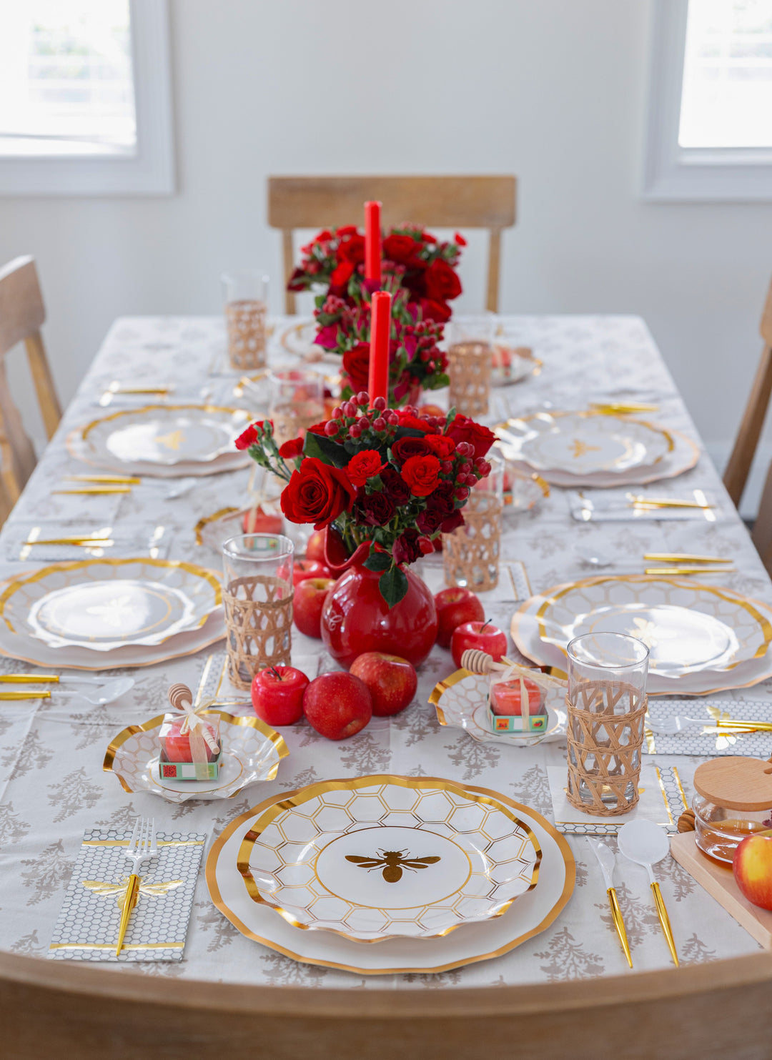 Sophistiplate LLC’s “Rosh Tablescape for 16 Guests: White Honeybee” sets a sophisticated Rosh Hashanah table with its gold-trimmed disposable plates featuring an elegant bee motif. Red apples, candles, and vases of red roses complement the gold cutlery and lattice-designed glasses, all artfully arranged around gift boxes on a patterned tablecloth.