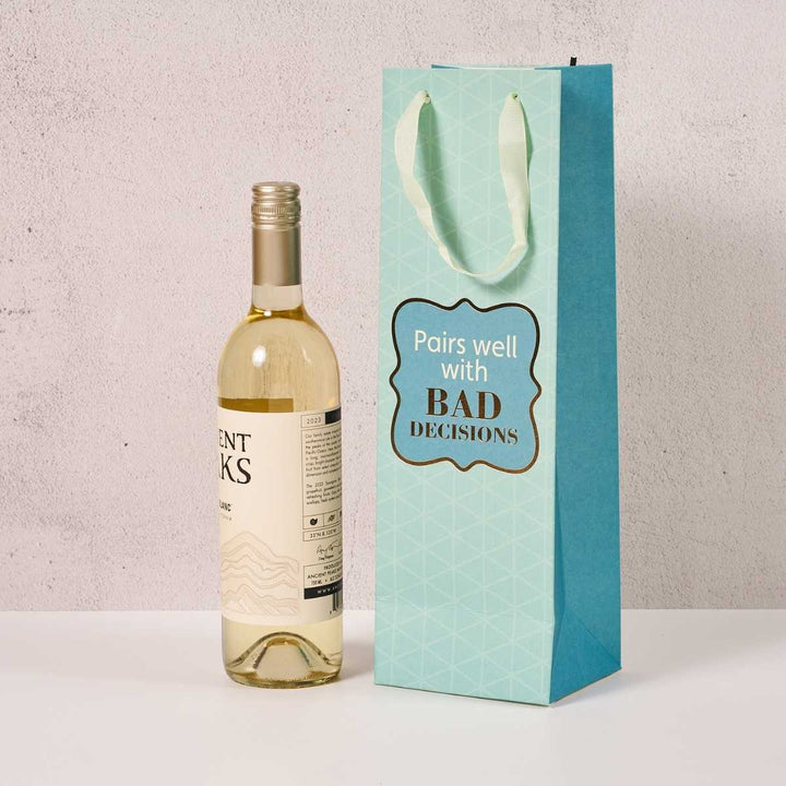 A bottle of white wine stands next to the Drinks On Me Bad Decisions-Wine Bag, featuring a light blue geometric design, pale handles, and the bold phrase "Pairs well with BAD DECISIONS"—a funny, ideal wine bag for any hostess gift.
