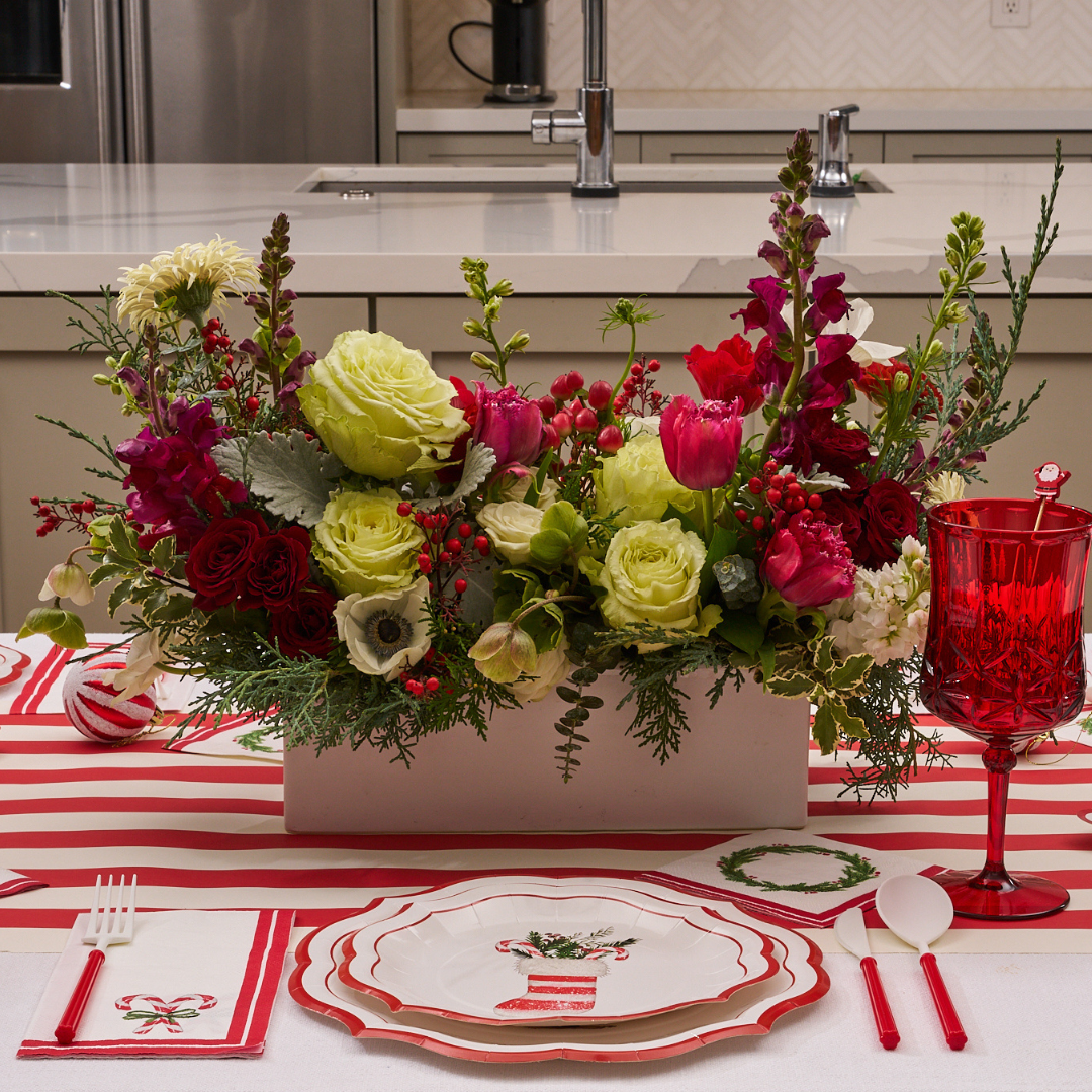 The Merry and Bright Table Setting by Sophistiplate features a festive striped runner, holiday plate, red glass, white cutlery, and a centerpiece with red, green, and white flowers—designer disposable plates complete the elegant display.