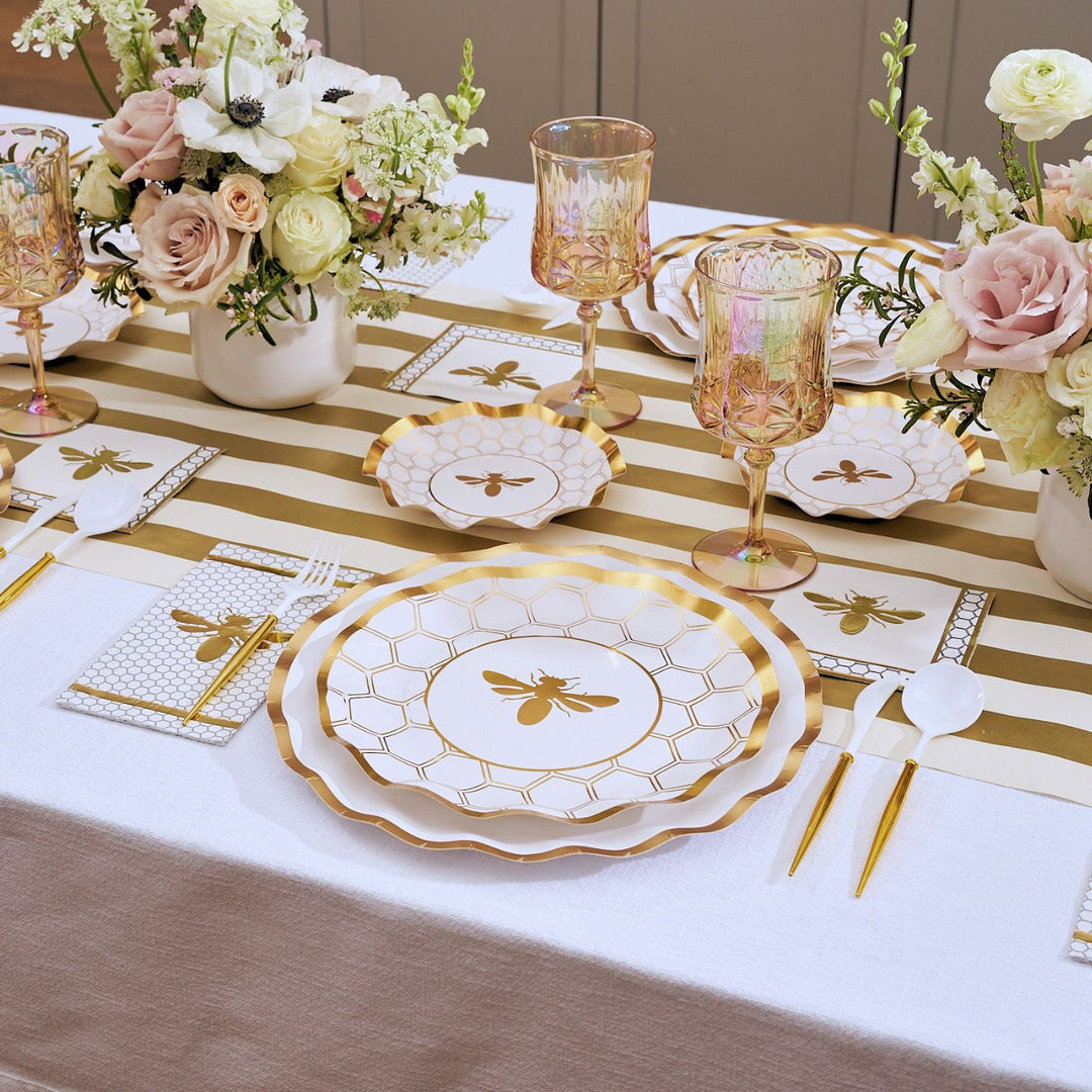 A table is set with the White Honeybee Table Setting by Sophistiplate, featuring bee-themed elegant party tableware such as gold rim paper plates, gold utensils, pink glasses, and floral centerpieces. The runner and napkins showcase gold stripes and bee designs for a coordinated look with stylish disposable plates—perfect for any celebration.