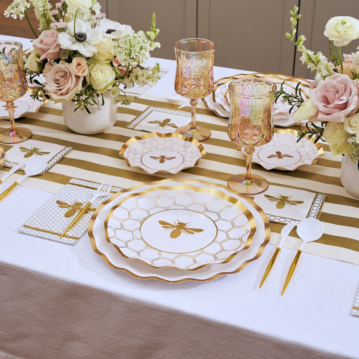 A table is set with the White Honeybee Table Setting by Sophistiplate, featuring bee-themed elegant party tableware such as gold rim paper plates, gold utensils, pink glasses, and floral centerpieces. The runner and napkins showcase gold stripes and bee designs for a coordinated look with stylish disposable plates—perfect for any celebration.